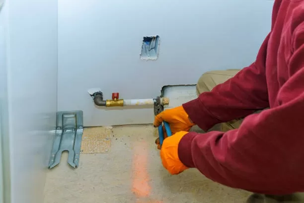 A plumber wearing orange safety gloves using a tool to connect a flexible gas line to a shut-off valve behind a kitchen cabinet.