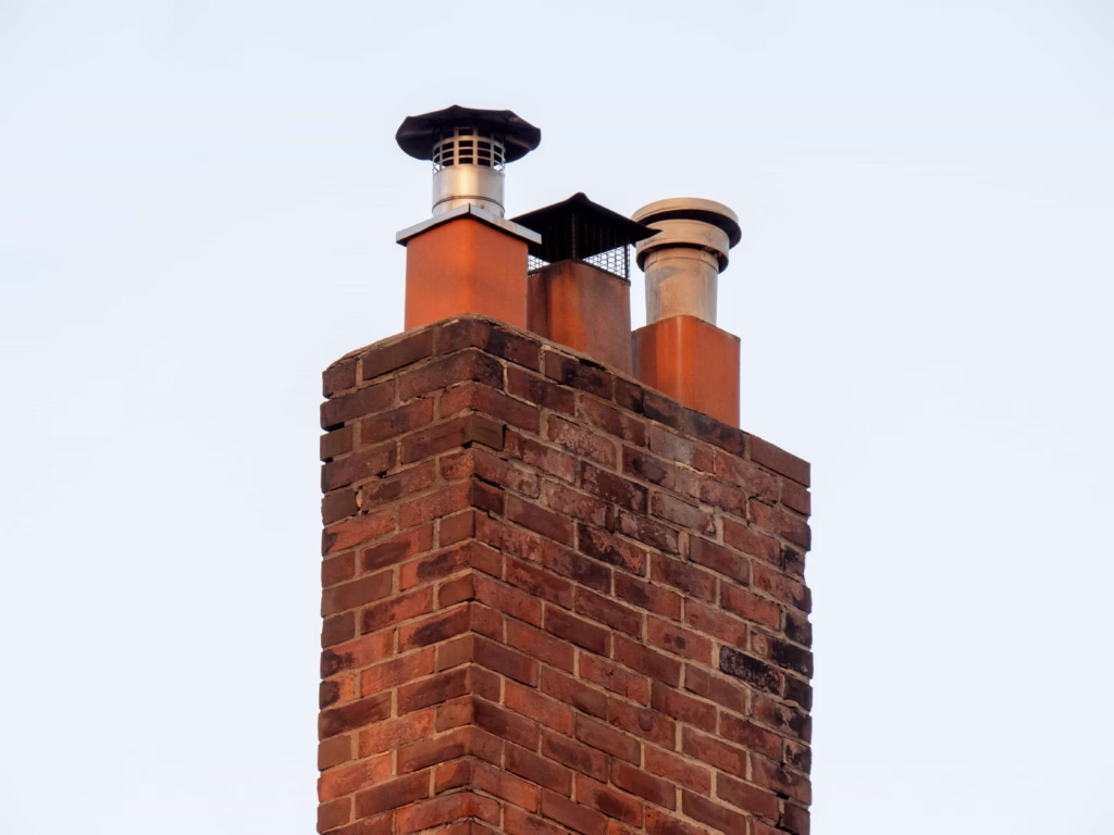 A red brick chimney with metal chimney caps against a light blue sky, representing expert chimney repair services in Boston, MA.