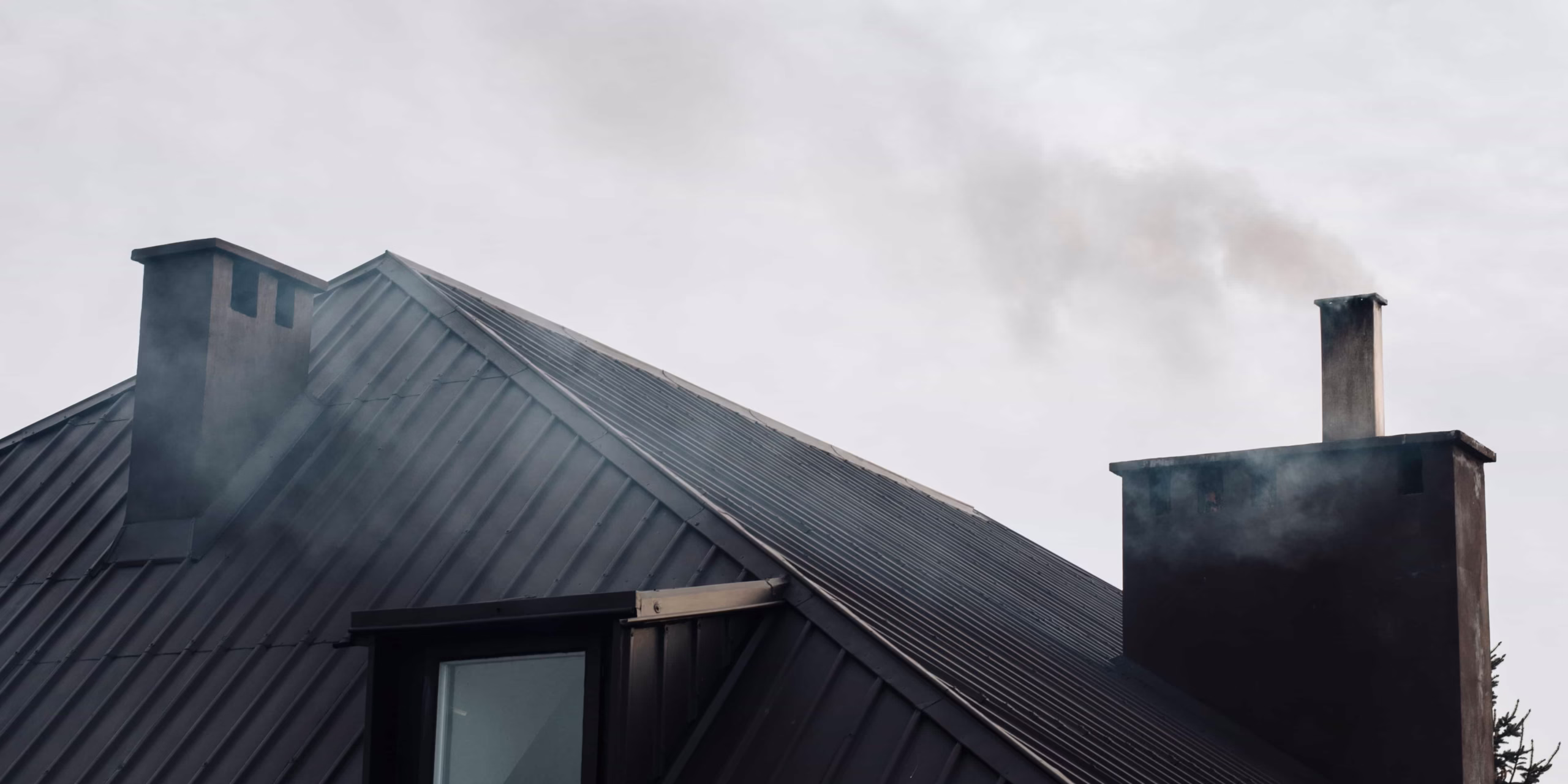 A residential dark metal roof with multiple chimneys releasing light smoke on a cloudy day, indicating active use that requires a professional to prevent a dormant chimney smell in Boston, MA.